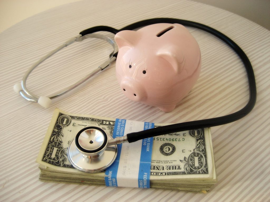 A stethoscope wrapped around a piggy bank and a wad of cash.